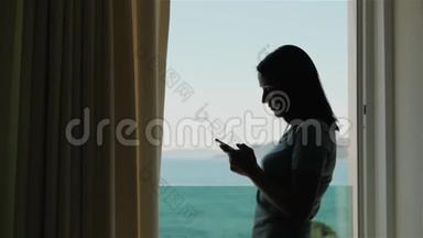 年轻女子智能手机剪影。 高加索模特站在窗口用她的智能手机
